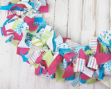 Add colorful style to your summer decorations with this handmade fabric garland banner. Hang it on a mantel, on the front of a dessert table, or drape it around along a stairwell for a dramatic statement piece. The possibilities are endless!
The garland is made using various raw-cut cotton fabric strips and ribbon that range between 8" and 10" long. They are then tied onto a thick strand of twine. T