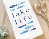 Blue 'Lake Life' kitchen towel  Form and function come together in this fun, flour-sack tea towel. Featuring a hand-drawn design, each towel has the phrase, "Lake Life," in dark blue letters. The phrase is surrounded by fish in light blue and dark blue shades. This would be a perfect gift for a lake enthusiast.   This item is proudly made in the USA.  Materials: Cotton  Dimensions: 27" wide x 27" long  Care: This item is machine washable.