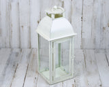 A white distressed candle lantern with a glass top and base, made of iron, displayed against a wooden backdrop.