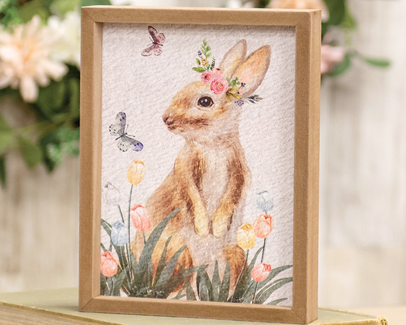 In a lovely mix of whimsy and rustic style, this small framed print will bring beauty to the farmhouse this spring. This piece features an inset, textured print and a natural wood frame. It depicts a bed of tulips in shades of blue, pink, yellow, and white with a brown and white bunny sitting up among them. 