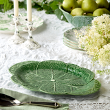 Green leaf-shaped ceramic plate on a table with white lace tablecloth, green apples, and white flowers.