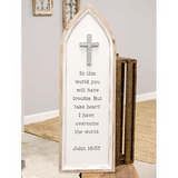 This wooden sign features a cathedral window design. It has a natural wood frame and cream background that reads, "In this world you will have trouble. But take heart! I have overcome the world -- John 16:33," in black lettering. It is accented by a cross at the top and comes ready to hang for immediate display. At almost 3 feet long this sign is perfect for a hallway or transition wall between a living room and kitchen.