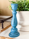 A distressed blue wooden candlestick holder with a flared base, standing on a white tray in a styled home setting with a potted green plant in the background.