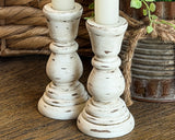 A pair of distressed white wooden candlestick holders with a scalloped, turned design, shown holding white candles on a rustic wooden surface with a wicker basket and metal pot in the background.