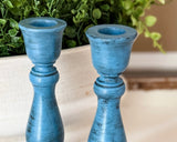 Two distressed blue wooden candlesticks with a rustic finish, shown in a close-up shot with green foliage in the background.