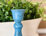 A close-up of a distressed blue candle holder or decorative stem holder with a flared top, shown in front of a blurred green plant and beige planter.