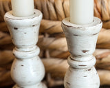 A close-up of two distressed white wooden candlestick holders with white candles, set against a warm, woven straw background.