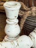 A close-up of a distressed white wooden candle holder with a turned spindle shape and a round top, shown next to a dark ribbed pot tied with jute rope on a wooden surface.