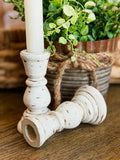 Two distressed white birch wood candlestick holders on a wooden table, with one holding a white candle and the other lying on its side. A potted green plant in a rustic metal pot sits in the background.
