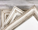 Three distressed white picture frames with rustic brown wood showing through the chipped paint, arranged diagonally on a gray wood-grain background.