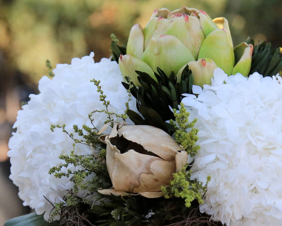 A striking single lifelike artichoke is nestled in an antique white, terra cotta pot and beautifully surrounded by preserved white hydrangea, greenery, concho pods, and natural grapevine accents. This elegant design blends classic charm with refined simplicity. At almost 1-foot tall, this arrangement is generously sized and perfect for adding a touch of nature-inspired beauty to any space.