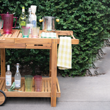 Wooden outdoor bar cart with drinks and glasses against a green bush background