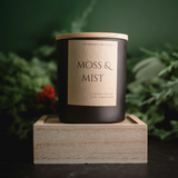 Candle labeled 'Moss & Mist' on a wooden block with a green foliage background