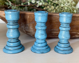 Three blue candle holders on a wooden surface with a wooden bowl and greenery in the background.