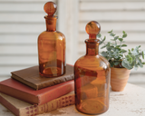 Set of 2 amber glass bottles with stoppers

Welcome vintage elegance with the unique and trendy design of these bottles. Display them as is or remove the matching glass top stopper to turn it into a stunning vase for your favorite flowers. This set of bottles is not food safe and is for decorative purposes only.&nbsp;

Your purchase includes two bottles, one of each size.

Materials:
Glass

Dimensions:
Smaller bottle -
2 1/2" diameter x 7" high

Larger bottle -
3" diameter x 8 1/2" high