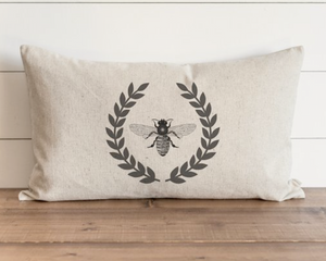 Beige pillow with bee and laurel wreath design on a wooden surface.