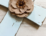 A decorative fabric flower with layered tan burlap petals and a brown center, placed on a distressed white wooden cross.