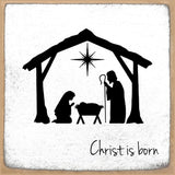 A square decorative sign or print with a nativity scene silhouette: a stable outline, star above, Mary kneeling on the left, Joseph standing on the right, and a baby in a manger. The phrase 'Christ is born' appears in black script near the bottom.