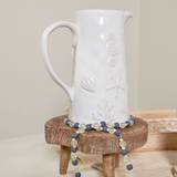 White ceramic pitcher with embossed designs on a wooden stand against a plain background