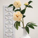 Three yellow flowers in a white vase against a light gray background with a decorative wall panel.