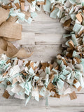  A full, handcrafted fabric rag wreath made of light robin's egg blue and white cotton strips on a round wire form, finished with a rustic burlap bow at the top.