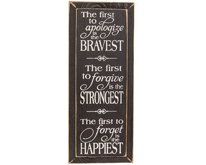 This sign is an inspirational wall art piece with two holes for versatile hanging display. Made from wood, the sign features a black stain that lets the natural grain peek through. A distressed finish gives the sign a rustic, farmhouse look. White text in a mix of script and print adorns the whole sign, accented by a few flourishes. The sign reads, “the first to apologize is the bravest, the first to forgive is the strongest, the first to forget is the happiest.” 