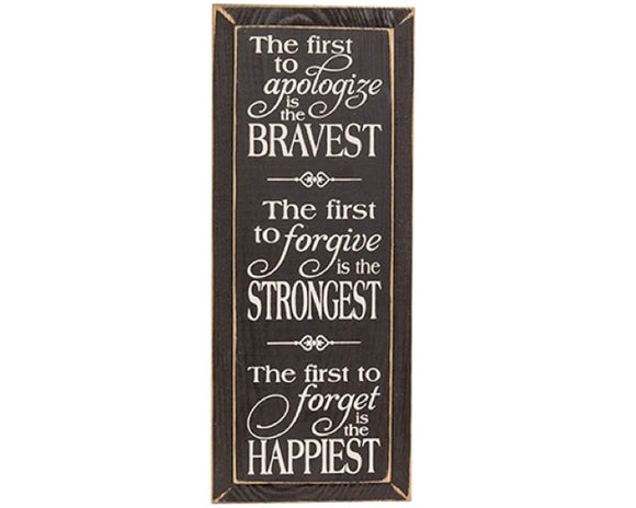 This sign is an inspirational wall art piece with two holes for versatile hanging display. Made from wood, the sign features a black stain that lets the natural grain peek through. A distressed finish gives the sign a rustic, farmhouse look. White text in a mix of script and print adorns the whole sign, accented by a few flourishes. The sign reads, “the first to apologize is the bravest, the first to forgive is the strongest, the first to forget is the happiest.” 
