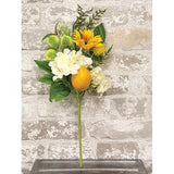 An artificial floral arrangement featuring white flowers, yellow sunflowers, green leaves, and a bright orange-yellow fruit, set against a distressed white brick wall background.