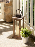 A rustic wooden lantern with a glass door and a black metal top handle, styled on a sunlit stone patio next to a potted green plant.