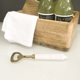 Two green bottles in a wooden holder with a white napkin and gold bottle opener on a white surface.