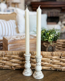 Two white pillar candles in distressed white ceramic candlestick holders, styled on a wooden surface in front of a woven basket with a blurred home interior background.