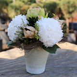 Floral arrangement with white flowers and green leaves in a textured pot on a wooden surface.