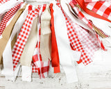 A rustic red, white, and beige fabric tassel garland made from gingham check, solid red, and solid white ribbons, hanging on a white distressed wooden surface.