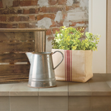 Metal pitcher and plant on a tiled surface with a brick wall background and red grain sack cube