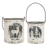 Two distressed white metal bucket lanterns with dark handles, featuring winter silhouettes of a deer on one and pine trees on the other, set against a plain white background.