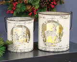 A nested pair of two white-painted, distressed metal farmhouse buckets featuring stenciled cutouts of a reindeer and a snowman, styled with winter greenery on a shelf.