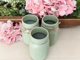 Three sage green painted mason jar candle holders tied with twine, arranged on a light wood surface with pink hydrangeas in the background.