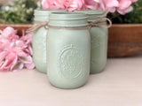 Three embossed mason jar candle holders in a muted sage green color, tied with twine, displayed on a light wooden surface with pink flowers in the background.