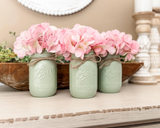 Three pale green Ball Mason glass jars tied with twine and filled with pink faux hydrangea flowers, displayed on a light wood surface in a rustic home decor setting.