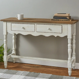 A distressed white console table with a rustic brown wooden top, featuring two drawers with round knobs, turned legs, and a lower support rail. It is styled in a room setting with a mug and books on top, set against a gray wall and wood floor.