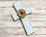 A rustic light blue wooden cross with distressed edges and a brown burlap flower accent at the center, displayed on a whitewashed wood plank background.