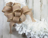 A close-up of a decorative bow made from natural burlap ribbon, placed on top of a white tinsel wreath against a whitewashed wood plank background.