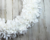 A white fabric wreath made of layered, ruffled paper-like strips, photographed against a distressed white wood plank background.