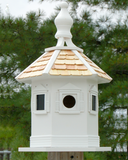 Decorative white birdhouse with a shingled roof against a green tree background