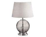 A coastal farmhouse table lamp featuring a round, smokey gray crackled glass orb base, a silver metal foundation, and a silvery neutral fabric drum shade.