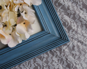 A large blue 16x20 wooden gallery photo frame with a black mat and a floral picture inside, displayed on a table with a lace tablecloth in the background.