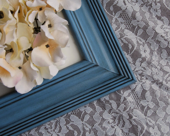 A large blue 16x20 wooden gallery photo frame with a black mat and a floral picture inside, displayed on a table with a lace tablecloth in the background.