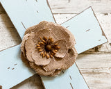 A spa blue and brown distressed wood wall cross with a burlap embellishment in the center, placed on a white wooden surface.