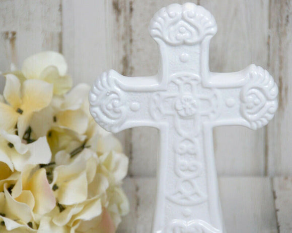 A white ceramic cross figurine with ornate detailing, displayed on a wooden surface with a floral decoration to the left.