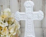 A white ceramic cross figurine with ornate detailing, displayed on a wooden surface with a floral decoration to the left.